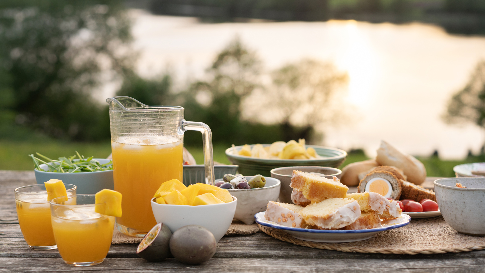 fresh fruit juice and picnic food on a picnic table with a lake behind
